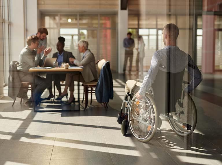 Rear view of a businessman in a wheelchair arriving on a meeting with his colleagues in the office. The view is through glass.