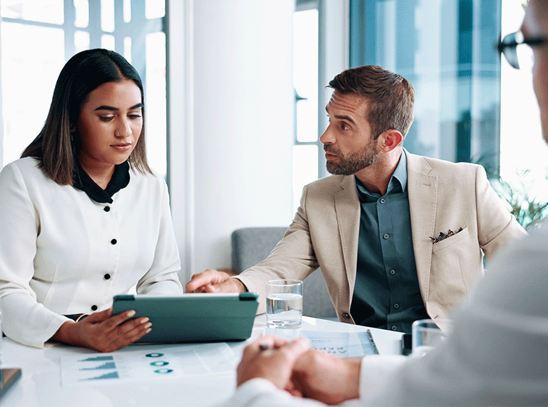 Business professionals in meeting, woman in white holding tablet, man in beige suit looking at colleague in bright office.