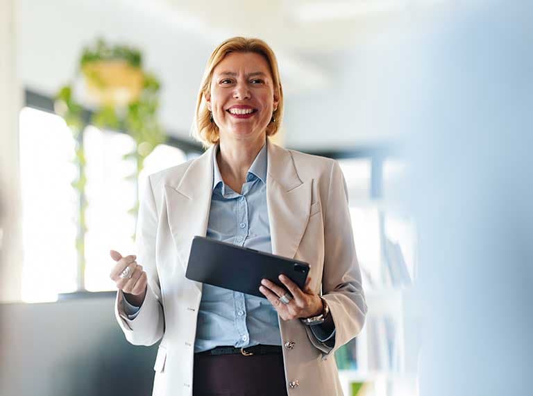Professional in white blazer and blue shirt smiling while holding a tablet in bright modern office with plants.