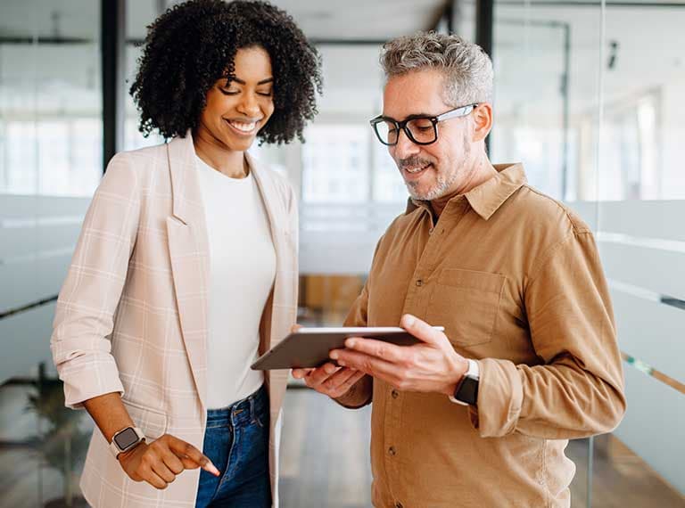 Two business professionals, a woman in a beige blazer and a man in glasses, reviewing content on a tablet in a modern office.
