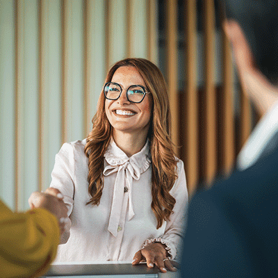 Woman with glasses and white blouse smiling while shaking hands with someone during a professional meeting or interview.