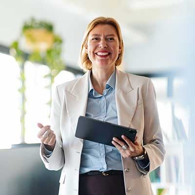 Professional in white blazer and blue shirt smiling while holding a tablet in bright modern office space.