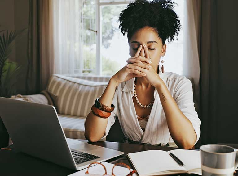Woman with natural hair rubbing her eyes while working at laptop in home office with notebook and coffee cup on desk.