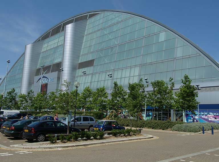 Xscape building in Milton Keynes - Snowdome, iFly. Modern building with curved glass and metal facade, parking lot with cars and trees in foreground under blue sky.