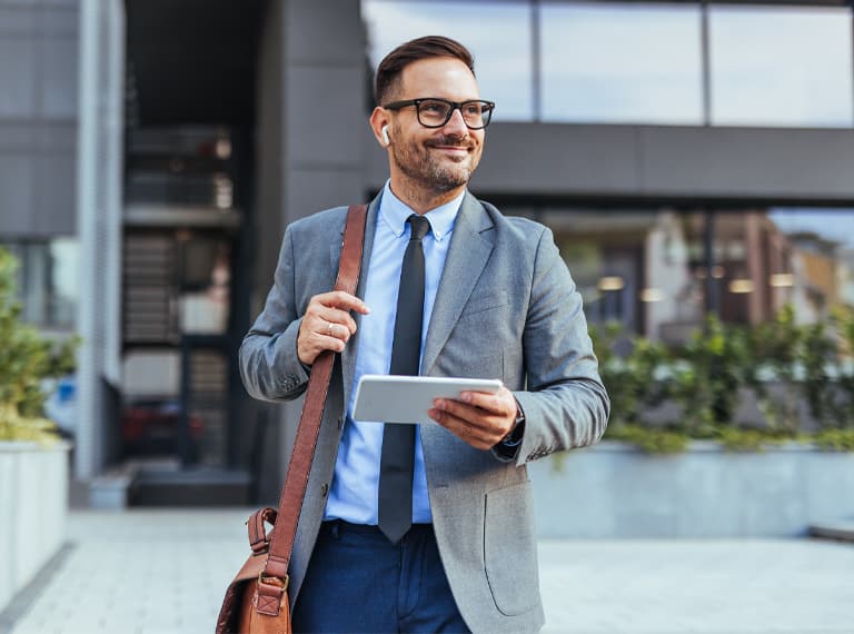 Smiling businessman in a suit and tie walking outside a modern office building, holding a tablet. He appears confident and professional, suggesting a successful business environment.