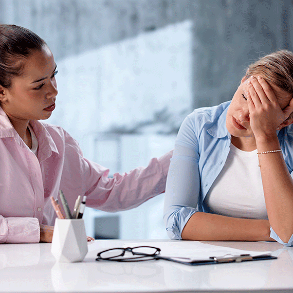 Woman in pink shirt comforting distressed colleague in blue shirt at office desk with glasses and phone visible.