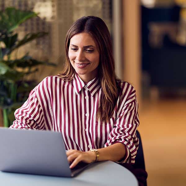 Person in red and white striped shirt smiling while working on laptop in modern office space with plants nearby.