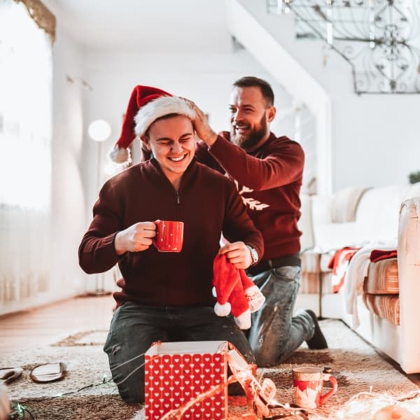 Smiling Gay Couple Opening Christmas Presents With Santa Hats And Drinking Tea Together
