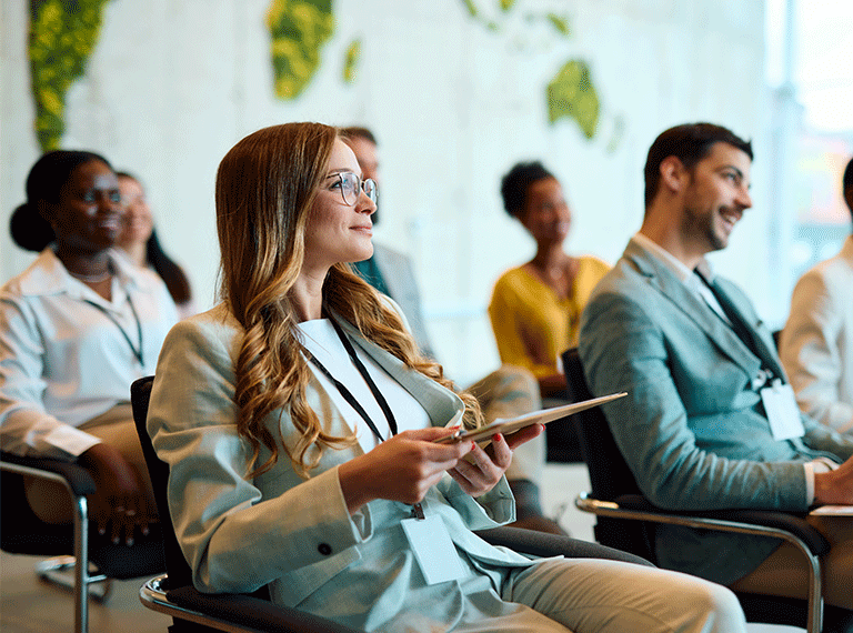 Diverse group of professionals sitting in a conference room, smiling woman with glasses holding tablet in foreground.