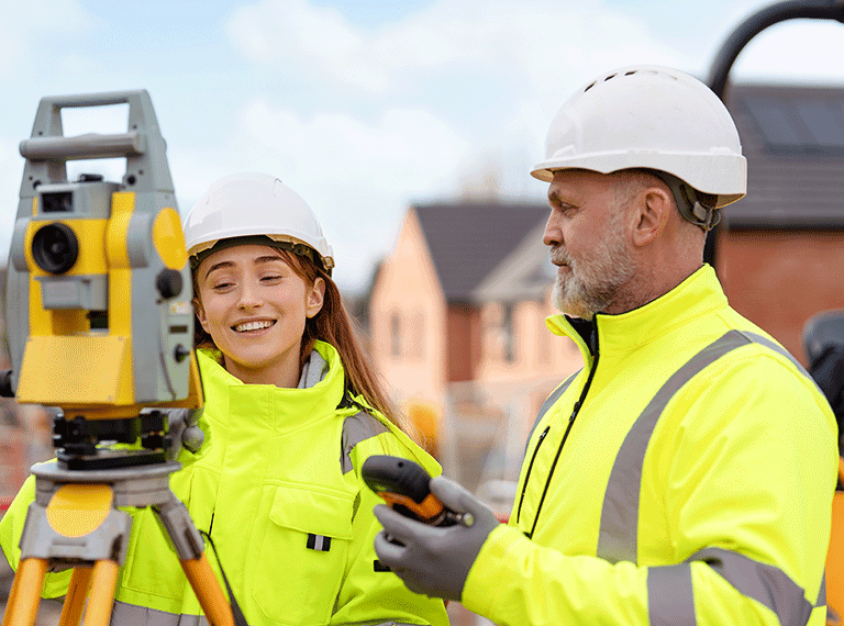 Two construction workers in white hard hats and yellow high-visibility vests at a job site with surveying equipment.