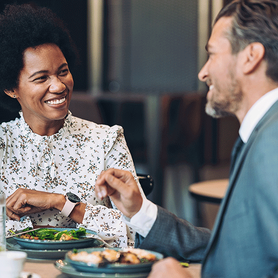 Woman with afro in floral blouse smiling at man in gray suit during business lunch with plates of food on table.