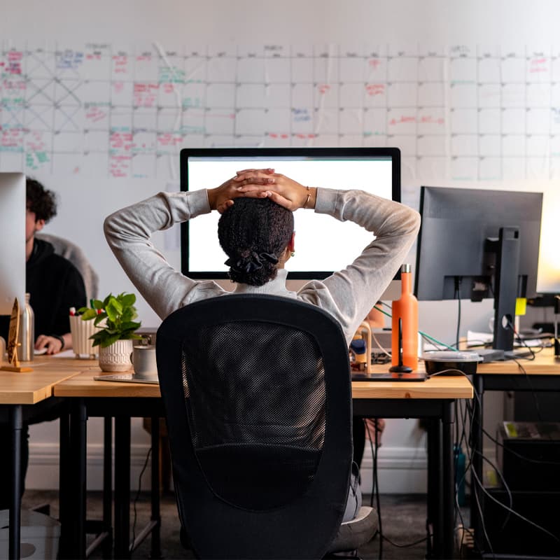 Rear view wide shot of a woman sitting working at a computer in an office. She has her hands behind her head as she stretches, looking at the screen.