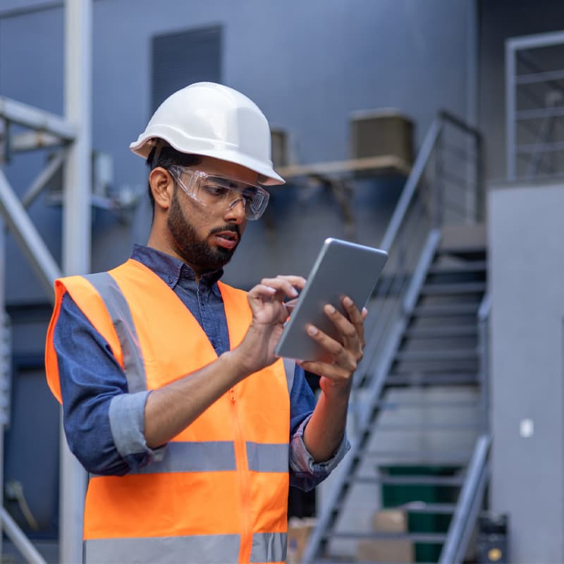 Serious thinking and focused engineer working in factory in hard hat and vest, man using tablet computer reading diagram, and checking equipment, inside industrial factory.