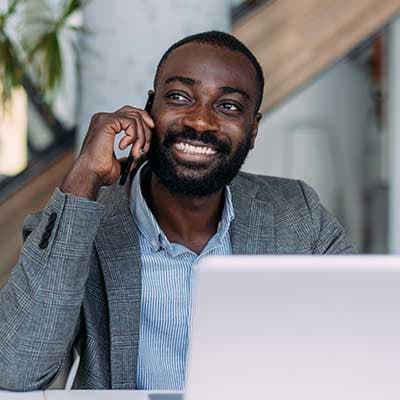 Smiling professional in gray blazer and striped shirt talking on phone while working on laptop in bright office space.