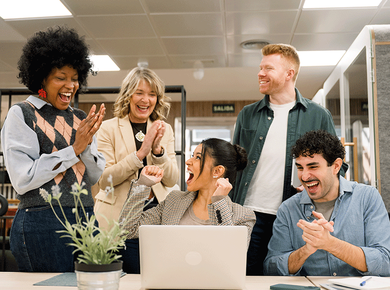 Five diverse coworkers celebrating together in a bright office, smiling and clapping around a laptop on a desk.