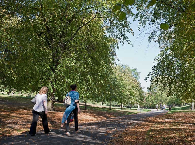 Two people walking on a tree-lined path in a park in Nottingham with green foliage and dappled sunlight on a clear day.