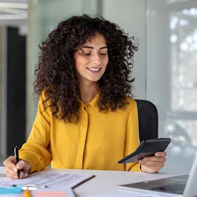 Person with curly hair in bright yellow blouse working at desk, holding phone while writing notes