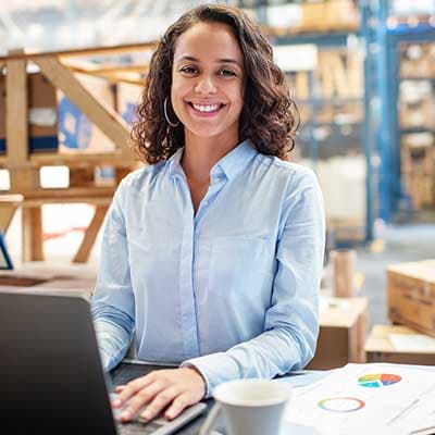 Smiling person in light blue shirt working on laptop in warehouse setting with coffee cup and charts nearby.