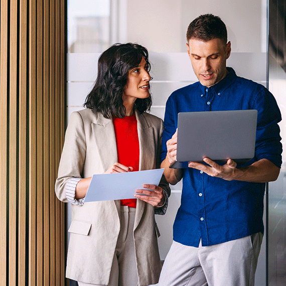 Two business professionals reviewing documents together, woman in beige blazer holding paper, man in blue shirt with laptop.