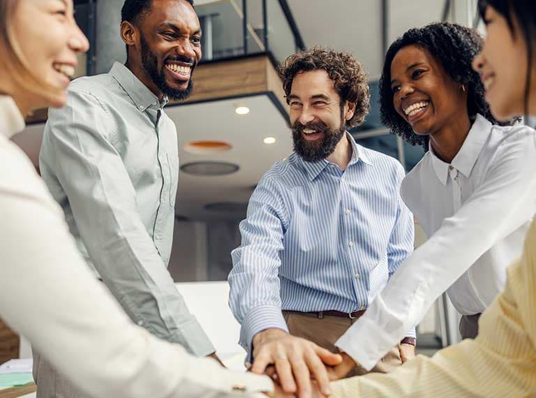Diverse group of smiling professionals joining hands together in a modern office setting, showing teamwork and unity.