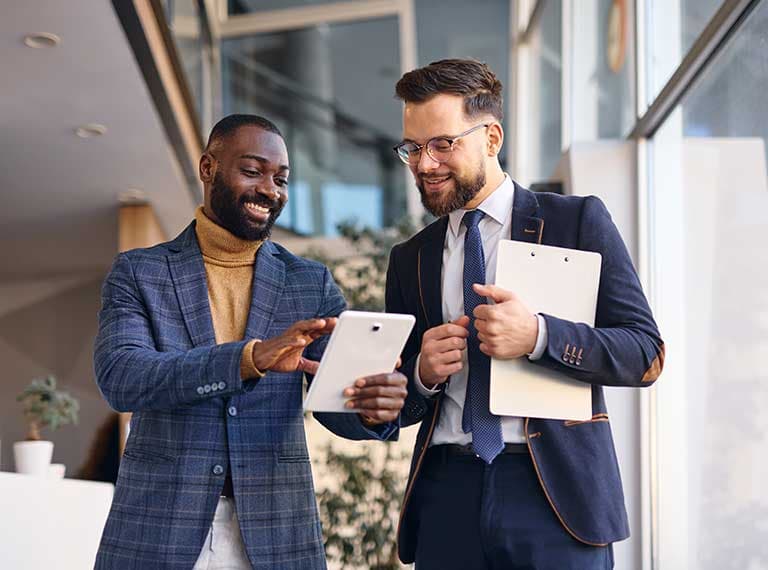 Two businessmen in suits smiling while looking at a tablet, one holding a clipboard in a modern office setting.