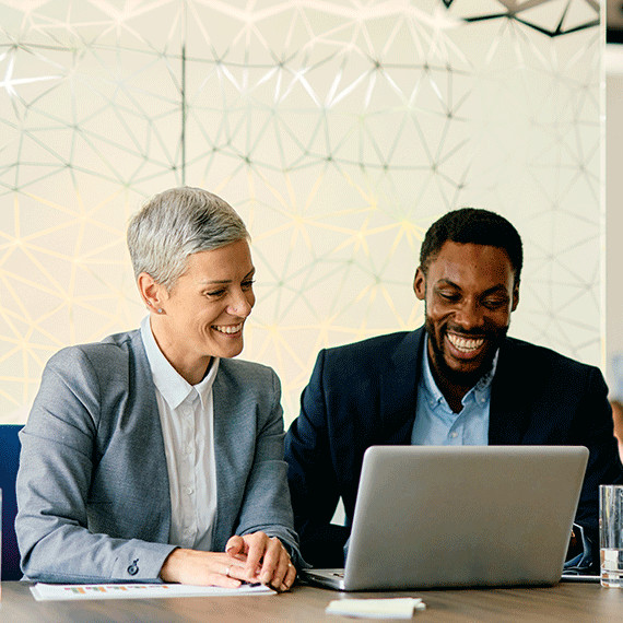 Two smiling business professionals in suits looking at a laptop together at a conference table with geometric wall art behind them.