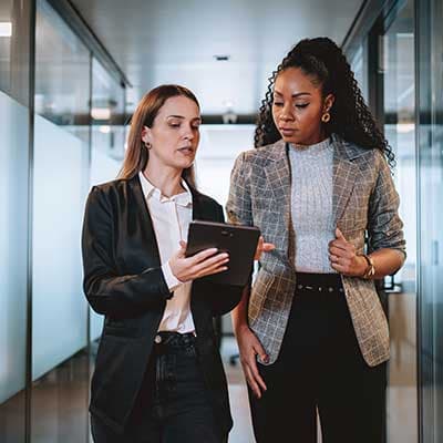 Two professionally dressed women reviewing information on a tablet while walking through a modern office corridor.